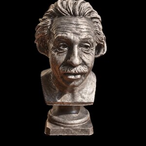 May include: A metallic silver-toned bust sculpture depicting the head and shoulders of Albert Einstein. The figure features detailed facial wrinkles, a prominent mustache, and textured hair, set upon a circular pedestal base.