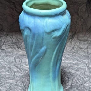 May include: A ceramic vase featuring a matte finish with a gradient of light turquoise to deep blue. The exterior surface displays vertical, raised, flowing relief patterns that extend from the base toward the circular, open rim at the top.