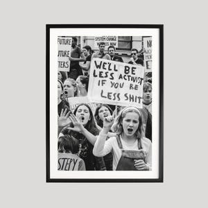 May include: A black and white framed photograph depicting a protest scene with multiple individuals holding signs. A prominent sign in the centre displays the text, "WE'LL BE LESS ACTIVIST IF YOU BE LESS SHIT," held by a person wearing overalls.