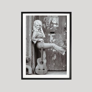 May include: A black and white photographic print featuring a person with voluminous blonde hair wearing a button-down shirt and jeans, leaning against a weathered door with the text DANCE ENSEMBLE, while holding an acoustic guitar.