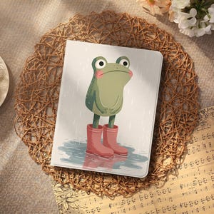 Puede incluir: Una funda rectangular para tableta o lector electrónico con una ilustración gráfica de una rana verde con mejillas rosadas de pie en un charco. La rana lleva botas de lluvia rojas y el fondo muestra una lluvia ligera sobre un fondo blanco.