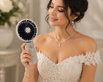 May include: A handheld portable fan featuring a dark navy blue circular head and a rectangular handle, both densely encrusted with clear, sparkling rhinestones. The center of the fan displays a decorative silver-toned floral embellishment.