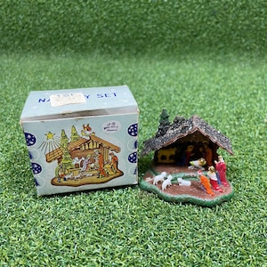 May include: A miniature nativity set featuring a small stable with a glittery roof and painted figurines of figures and animals, accompanied by a light blue cardboard box labeled NATIVITY SET with a printed illustration of the scene.