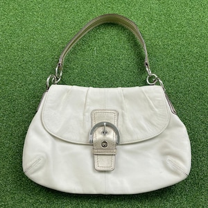 May include: A white leather shoulder bag featuring a single handle, a fold-over flap, and a central metallic buckle closure. The bag has a soft, rounded silhouette with subtle pleating details near the top and silver-toned hardware accents.