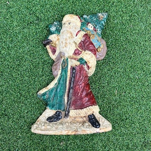 May include: A vintage-style cast metal Santa Claus figurine featuring a red and green coat with white trim, a long white beard, and a black walking stick. Santa carries a large sack filled with toys and a Christmas tree on his back.
