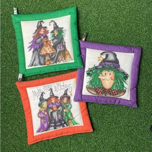 May include: Three square fabric pot holders with hanging loops. One has a green border and two witches, one has a purple border and a single witch face, and one has an orange border with three witches and the text Make three Witches.