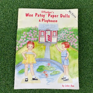 May include: A paper doll book titled Effanbee's Wee Patsy Paper Dolls & Playhouse, Wee Edition by John Axe. The cover features a watercolor illustration of two children standing by a pond with a small house in the background and pink flowers.