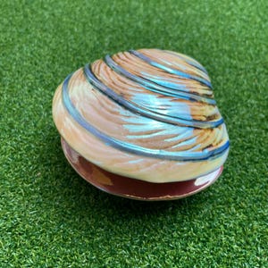 May include: A ceramic clam shell trinket box featuring a glossy finish with a cream-colored base and iridescent blue ridges. The lid is textured with raised, curved lines that follow the natural shape of the shell, creating a reflective surface.