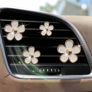 Può includere: Quattro clip per bocchette d'aria dell'auto a forma di fiore bianco con bordi dorati e un perno centrale dorato. I fiori sono fissati alle lamelle nere orizzontali di una bocchetta di ventilazione, con una finitura lucida ed effetti luminosi.
