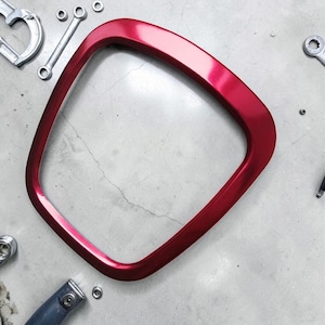 Könnte beinhalten: Ein glänzender, metallisch roter, abgerundeter quadratischer Rahmen mit einem glatten, geschwungenen Rand. Der Rahmen ist von verschiedenen silberfarbenen Metallwerkzeugen umgeben, darunter Schraubenschlüssel und eine Zwinge, die auf der hellgrauen Oberfläche verstreut sind.