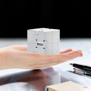 Puede incluir: Un cubo infinito metálico de color plateado compuesto por ocho cubos más pequeños conectados por bisagras. El objeto presenta una superficie lisa y reflectante, construida con juntas metálicas visibles que permiten plegar y desplegar el cubo.