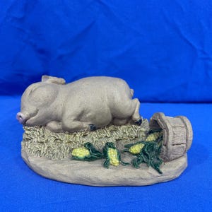 May include: A tan pig figurine lying on a textured base resembling hay. The pig has a pink snout and black hooves. Next to the pig is a small, overturned wooden bucket with several ears of yellow corn with green husks scattered nearby.