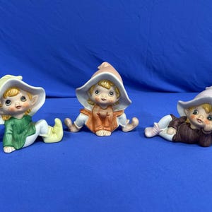 May include: Three ceramic elf figurines with blonde hair and pointed hats. The left elf wears a green tunic, the center elf wears a brown tunic, and the right elf wears a dark brown tunic while reclining. Each figure has painted facial features.