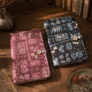 May include: Two rectangular fabric-covered journals, one in deep magenta and one in dark blue, each featuring a patchwork pattern of small illustrations including cats, rabbits, and floral motifs. Both journals are secured with a single light-coloured button.