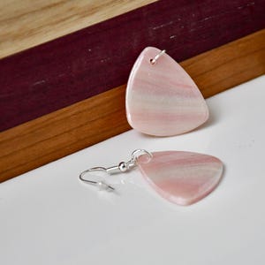 Könnte beinhalten: Ein Paar Ohrringe mit glatten, dreieckigen rosa Steinanhängern mit feiner weißer Maserung. Ein Anhänger ist an einem silberfarbenen Metallhaken befestigt, während der andere flach liegt und eine polierte, glänzende Oberfläche mit abgerundeten Ecken zeigt.