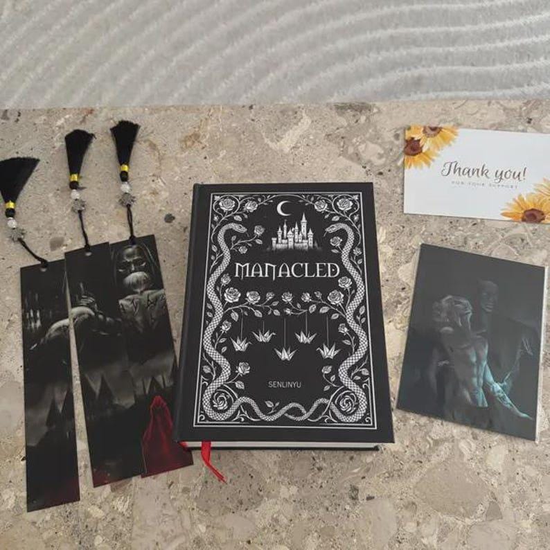 May include: A black hardcover book titled Manacled by Senlinyu with white cover art featuring a castle, roses, snakes, and origami cranes. Three black bookmarks with tassels and a black-and-white illustration card are included with a thank you note.