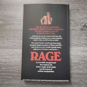 Puede incluir: Una portada de libro de bolsillo negra con el t&iacute;tulo RAGE en letras rojas grandes. La portada presenta una peque&ntilde;a fotograf&iacute;a de una persona en la parte superior y texto blanco que describe una historia sobre Charlie y un juego peligroso.