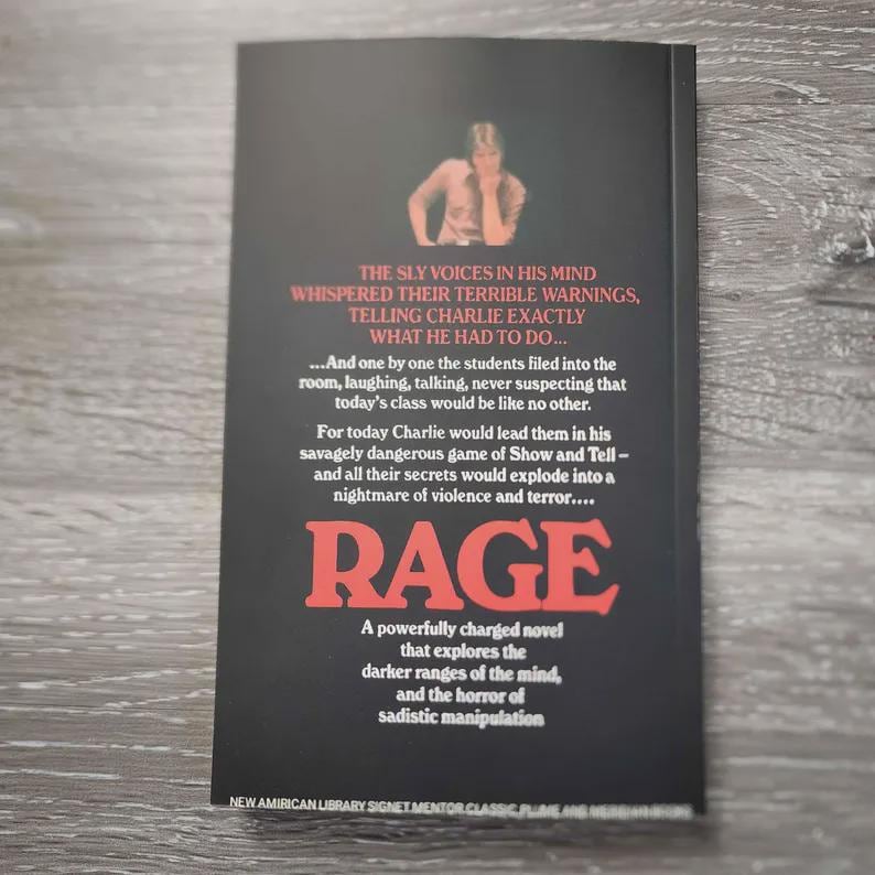 Puede incluir: Una portada de libro de bolsillo negra con el t&iacute;tulo RAGE en letras rojas grandes. La portada presenta una peque&ntilde;a fotograf&iacute;a de una persona en la parte superior y texto blanco que describe una historia sobre Charlie y un juego peligroso.