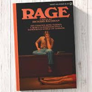Puede incluir: Un libro de bolsillo titulado RAGE de Richard Bachman con una cubierta negra y texto grande en naranja. La ilustraci&oacute;n muestra a una persona sentada en un escritorio en una habitaci&oacute;n oscura, con el texto: HIS TWISTED MIND TURNED A QUIET CLASSROOM INTO A DANGEROUS WORLD OF TERROR.