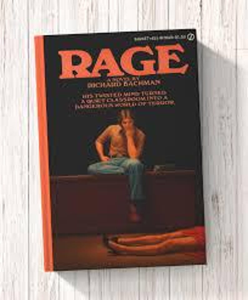 Puede incluir: Un libro de bolsillo titulado RAGE de Richard Bachman con una cubierta negra y texto grande en naranja. La ilustraci&oacute;n muestra a una persona sentada en un escritorio en una habitaci&oacute;n oscura, con el texto: HIS TWISTED MIND TURNED A QUIET CLASSROOM INTO A DANGEROUS WORLD OF TERROR.