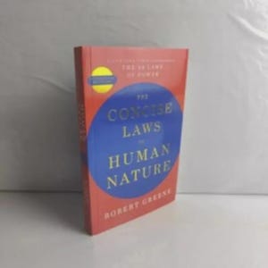 K&ouml;nnte beinhalten: Ein gebundenes Buch mit dem Titel The Concise Laws of Human Nature von Robert Greene. Das Cover zeigt einen gro&szlig;en blauen Kreis auf rotem Hintergrund mit goldenem Text. Ein gelber, runder Aufkleber befindet sich oben links auf dem Cover.