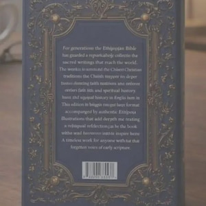 May include: A dark blue hardcover book featuring an ornate gold-colored filigree border on the back cover. The center contains printed text about the Ethiopian Bible and a barcode at the bottom. The design includes intricate scrollwork patterns.