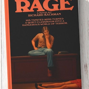 Puede incluir: Una portada de libro titulada RAGE de Richard Bachman con texto naranja sobre fondo negro. La ilustraci&oacute;n muestra a una persona sentada en un escritorio, vistiendo una camisa marr&oacute;n y pantalones azules, con el texto: HIS TWISTED MIND TURNED A QUIET CLASSROOM INTO A DANGEROUS WORLD OF TERROR.