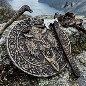 May include: A circular wooden shield featuring a carved wolf head at the center, surrounded by intricate knotwork patterns. A metal-headed axe with a decorated wooden handle rests across the shield, accompanied by a carved wooden staff.