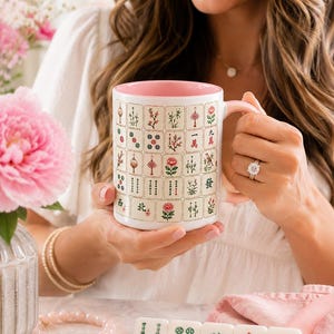 Peut inclure: Une tasse en c&eacute;ramique blanche avec un int&eacute;rieur et une anse roses, orn&eacute;e d'un motif r&eacute;p&eacute;titif de tuiles de Mahjong color&eacute;es incluant des fleurs, des caract&egrave;res chinois et des symboles circulaires. La tasse est tenue par une personne portant une bague.