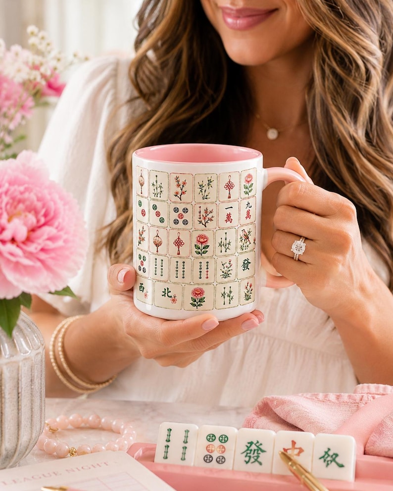 Peut inclure: Une tasse en c&eacute;ramique blanche avec un int&eacute;rieur et une anse roses, orn&eacute;e d'un motif r&eacute;p&eacute;titif de tuiles de Mahjong color&eacute;es incluant des fleurs, des caract&egrave;res chinois et des symboles circulaires. La tasse est tenue par une personne portant une bague.