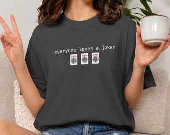 May include: A dark gray t-shirt featuring the white text "everyone loves a joker" printed above three small playing card graphics, each depicting a joker card with a central circular design and the word "JOKER" at the top of each card.