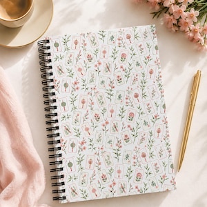 Puede incluir: Un cuaderno de espiral con una cubierta blanca que presenta un patrón repetitivo de fichas de mahjong, ramas de flores rosas y pequeñas linternas rojas. Las fichas muestran caracteres chinos verdes y detalles en rojo sobre un fondo floral.