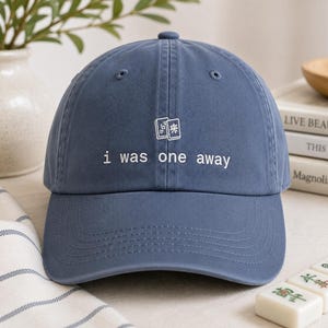 May include: A dusty blue baseball cap featuring white embroidered text that reads i was one away, positioned below a small white graphic of two mahjong tiles. The cap has a structured crown, a curved brim, and stitched eyelets on the top panels.