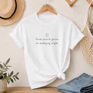 Könnte beinhalten: Ein weißes kurzärmeliges T-Shirt mit einer kleinen Grafik eines Mahjong-Steins über dem schwarzen Text from match point to mahjong night. Das T-Shirt hat einen Rundhalsausschnitt und ist auf einem Holzkleiderbügel mit geknotetem Saum.