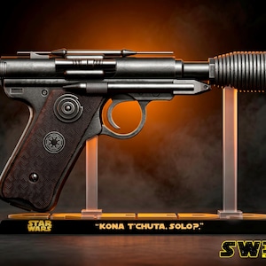 Può includere: Una replica in scala 1:1 di una pistola blaster con finitura metallica, dotata di un'impugnatura testurizzata marrone scuro con un emblema imperiale. Il blaster è montato su un supporto nero con il testo "STAR WARS" e "KONA T'CHUTA, SOLO?."