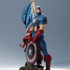 May include: A Captain America figurine standing on a rocky base, wearing a blue textured suit with a white star on the chest, red gloves, and red boots. The figure holds a circular red, white, and blue shield and a large American flag on a gold pole.