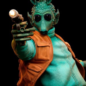May include: A green alien figure with a textured, bumpy head and large black eyes, wearing a teal zip-up jacket and an orange utility vest with pockets. The figure holds a metallic blaster pistol with a glowing light at the end of the barrel.