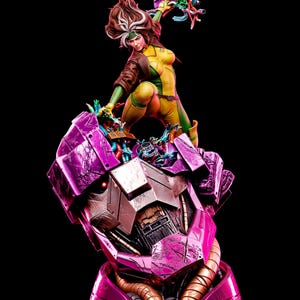 May include: A detailed collectible statue featuring a character with brown hair and a white streak, wearing a yellow and green bodysuit with a brown jacket, standing atop a large, damaged, purple and metallic robotic head with glowing red eyes.