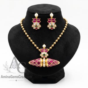 May include: A gold-toned necklace and earring set featuring deep pink, faceted, diamond-shaped stones arranged in a lattice pattern. The necklace includes a central pendant with a pearl accent, and the earrings match the pendant's floral design.