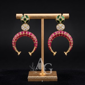 May include: A pair of gold-tone earrings featuring a crescent-shaped base inlaid with small red square stones. Above the crescent, a circular section contains four white stones, topped by a round green stone set in a gold frame.