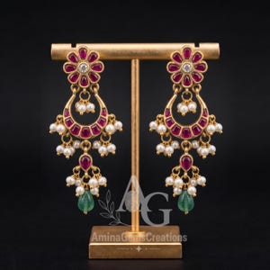 May include: A pair of gold-toned dangle earrings featuring a flower-shaped top with a central clear stone, a crescent-shaped middle section with inset magenta stones, and dangling white pearls with a single green teardrop stone at the base.