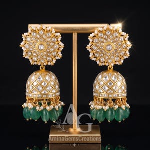 May include: A pair of gold-toned jhumka earrings featuring a circular floral top with inset clear stones and a bell-shaped base with a lattice pattern, accented by small white beads and dangling oval-shaped green stones, displayed on a gold stand.