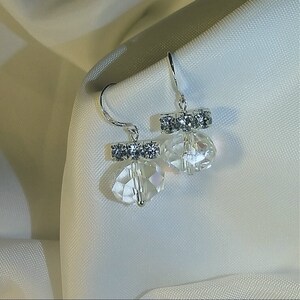 Puede incluir: Un par de elegantes pendientes colgantes. Cada pendiente presenta un gancho plateado, una barra con cristales brillantes y un colgante de cristal transparente facetado. Los pendientes se presentan sobre un fondo blanco suave, resaltando su delicado diseño.