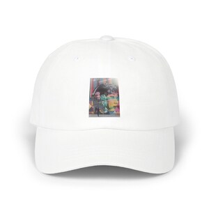 May include: A solid white baseball cap featuring a rectangular graphic print on the front panel. The print displays a colorful mural with a stylized face, pink floral elements, and a dark silhouette figure against a vibrant, multi-colored background.