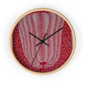 May include: A round wall clock with a light wood frame and black hands. The clock face features a white pumpkin graphic covered in red polka dots, set against a vibrant red background with a white, intricate, organic, net-like pattern.