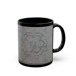 May include: A black ceramic mug featuring a central gray panel with a textured, stone-like appearance. The panel displays a geometric, star-shaped floral pattern in a lighter gray tone, centered on the front of the mug with a black handle.