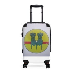 May include: A white rolling suitcase featuring a central circular graphic on the front. The graphic displays two stylized teal dogs standing side-by-side against a yellow background, bisected by a horizontal red stripe and a small central flower.