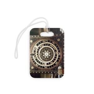 May include: A rectangular luggage tag featuring a black and gold circular astrological design with intricate patterns and small animal icons. The tag includes a white plastic loop at the top for attachment, showcasing a detailed celestial-inspired motif.