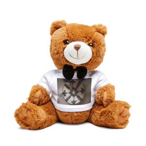 Puede incluir: Un oso de peluche marrón con hocico blanco y nariz marrón oscuro. El oso lleva una pajarita negra y una camiseta blanca con un estampado gráfico rectangular de un plato de cerámica circular con motivos azules y blancos y una estrella.