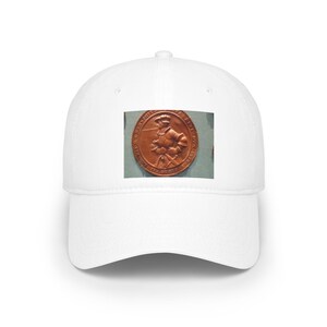 May include: A white baseball cap featuring a centered rectangular graphic. The graphic displays a circular copper-toned medallion with a relief image of a figure in historical armor, surrounded by circular text inscriptions along the outer edge.